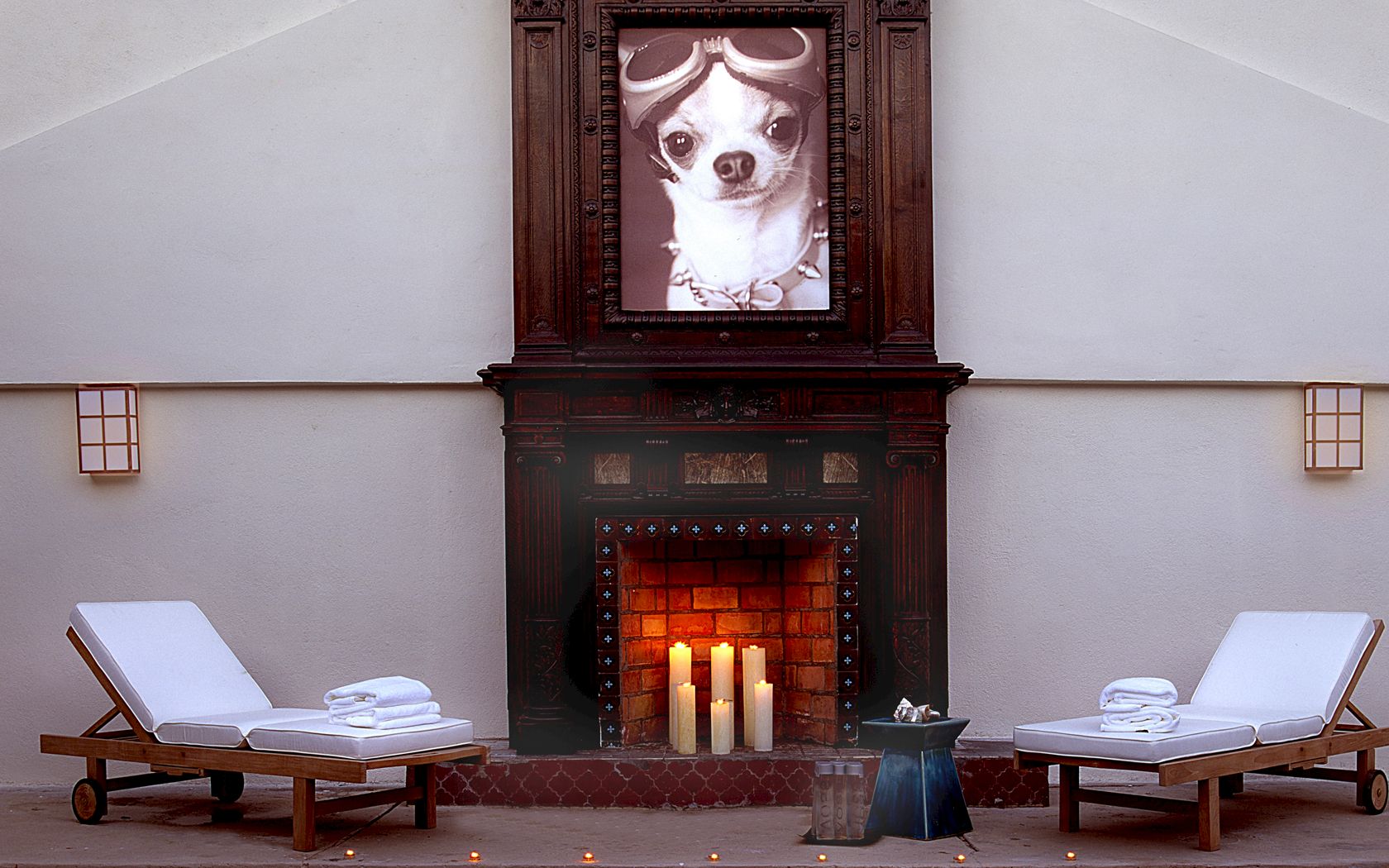 A cozy spa-like patio with two lounge chairs, fluffy towels, and a lit fireplace framed by a creepy portrait of a dog in a dark mantel.