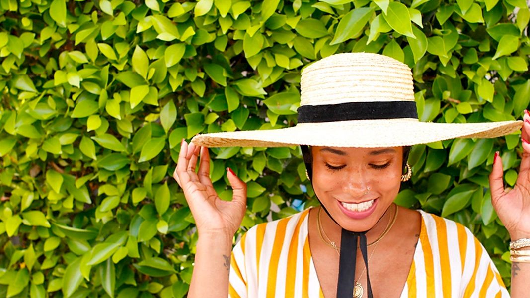 A smiling woman wearing a striped yellow and white outfit and a wide-brimmed hat stands in front of a lush green hedge, holding her hat's brim.