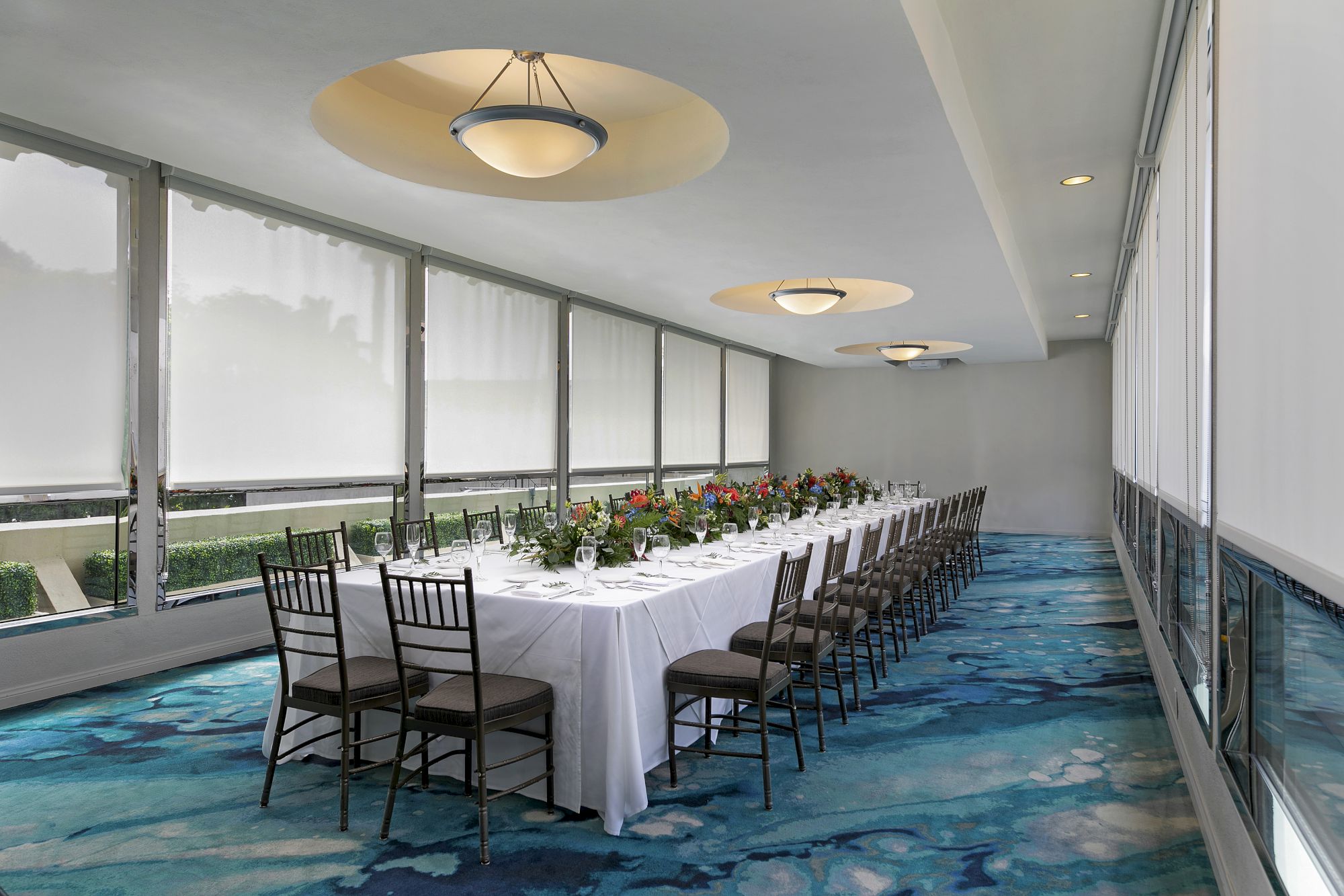 An elegant banquet setup with white tablecloth, glassware, floral centerpieces, and pendant lights in a sunlit room.