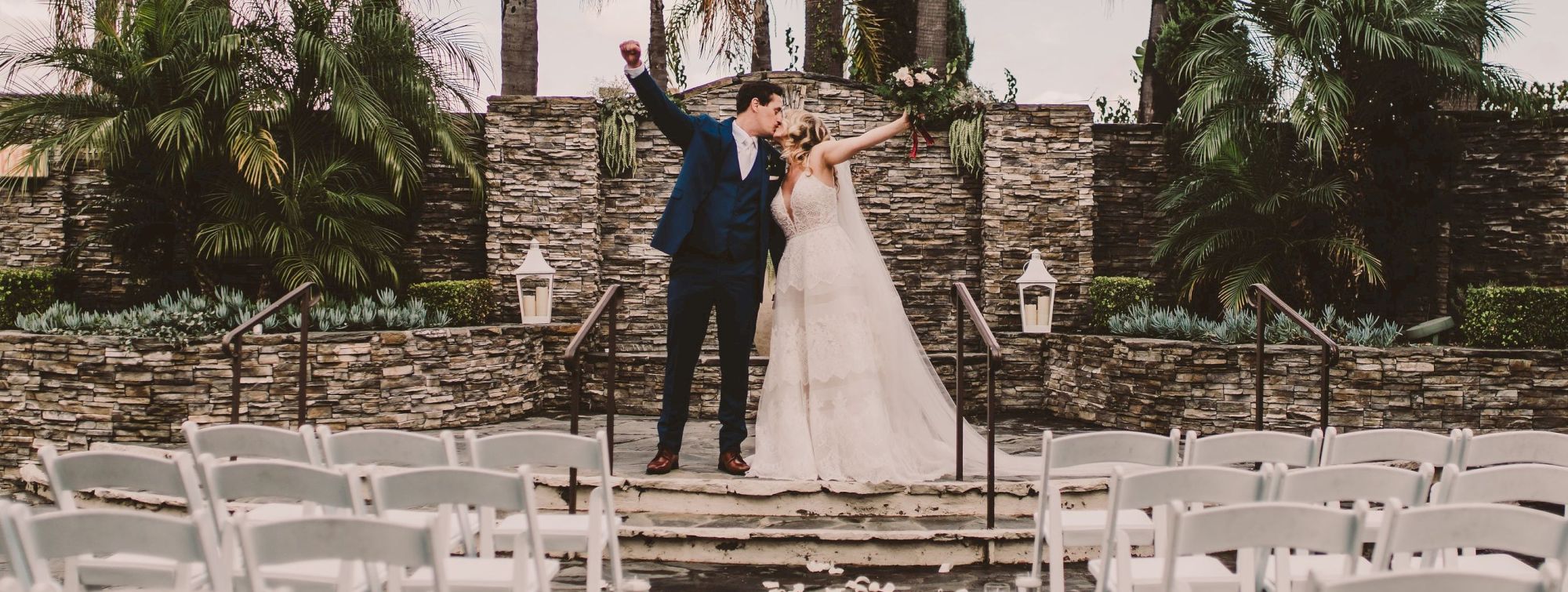 A bride and groom pose on an outdoor wedding stage, surrounded by palm trees and stone walls, with lantern decor nearby.
