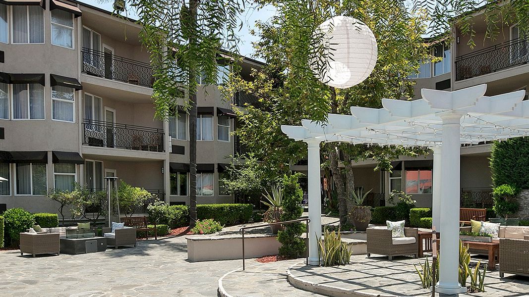 A sunny hotel courtyard with a white pergola, outdoor seating, round paper lanterns, umbrellas, and greenery in a multi-story building backdrop.