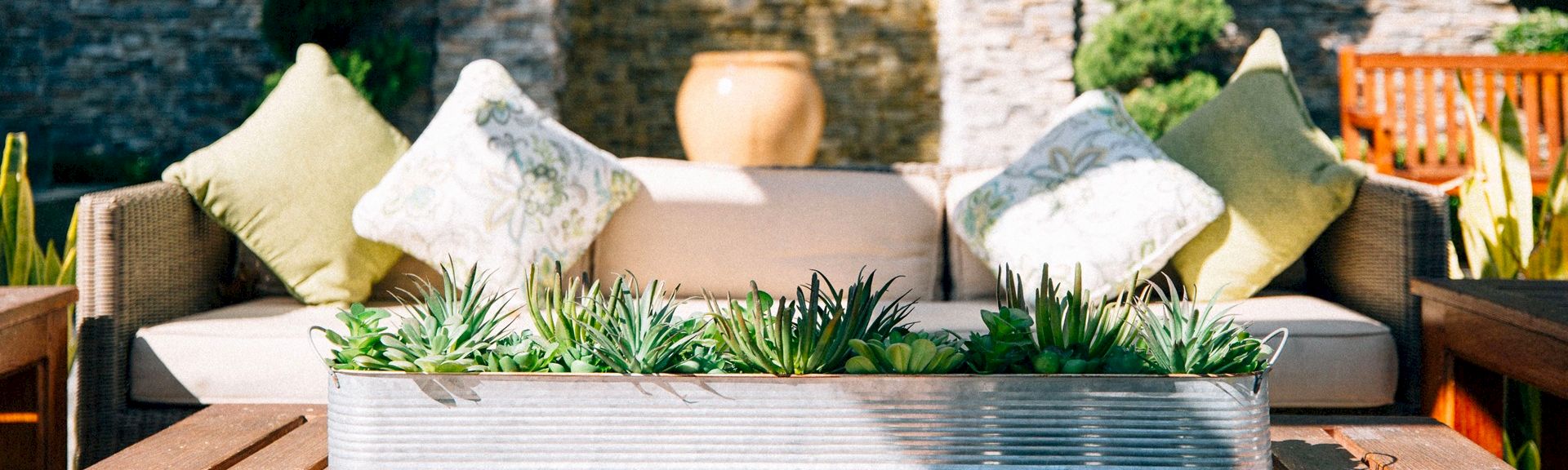 Outdoor patio sofa with decorative pillows and a metal planter filled with green succulents on a sunny day.