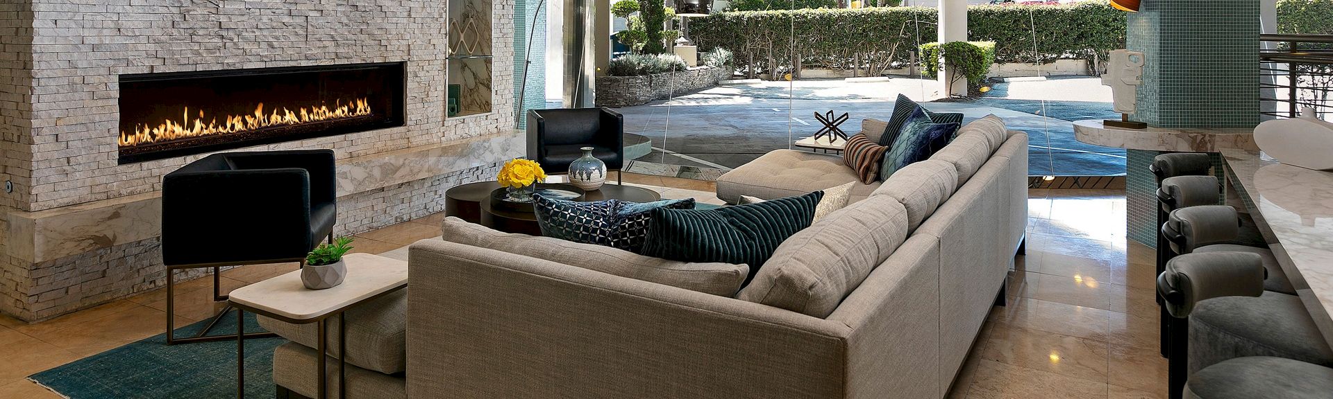 A luxury patio lounge with a roaring indoor fireplace, beige sectional sofas, and a poolside courtyard with greenery.
