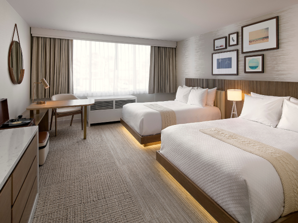 A modern hotel room with two queen beds, a desk and chair, a flat-screen TV, warm lighting, and framed art on the wall, bright window in the background.