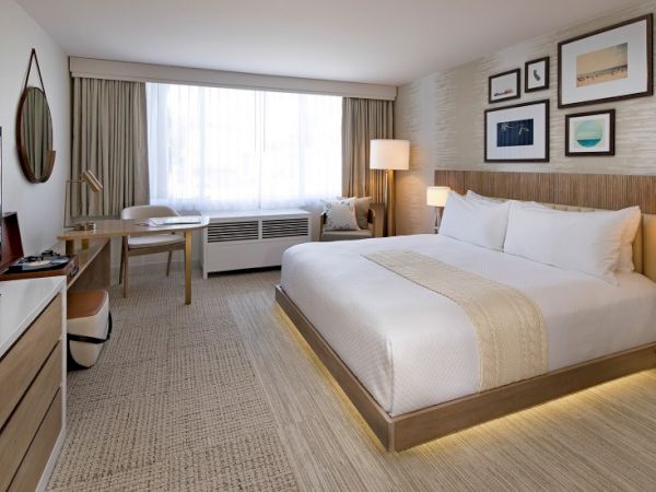 A modern hotel room with a large bed, desk, sofa, and framed wall art, bright natural light, and warm neutral tones. End sentence.