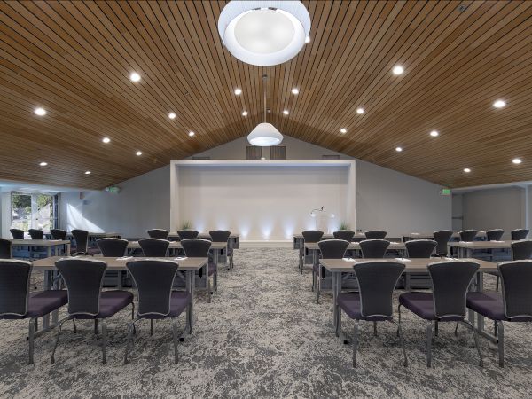 A bright conference room with rows of desks and chairs, a lectern at the front, and recessed ceiling lights.