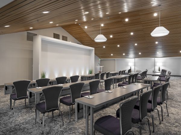 A modern conference room with long tables, cushioned chairs, pendant lights, and a wood-paneled ceiling.