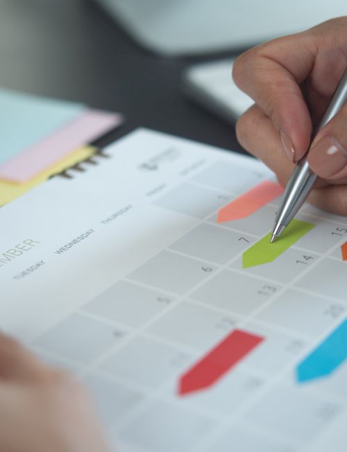 A hand marks a colorful calendar page with a pen, planning dates and schedules.