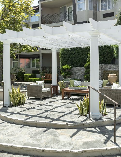 Sunny backyard patio with a white pergola, string lights, stone pavers, and cushioned outdoor seating among lush greenery.