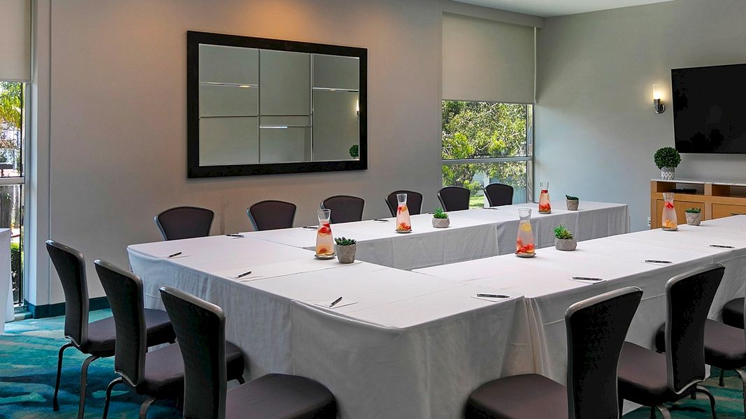 A modern meeting room with a long table, chairs, small plants, and a window-side buffet of food and drinks.