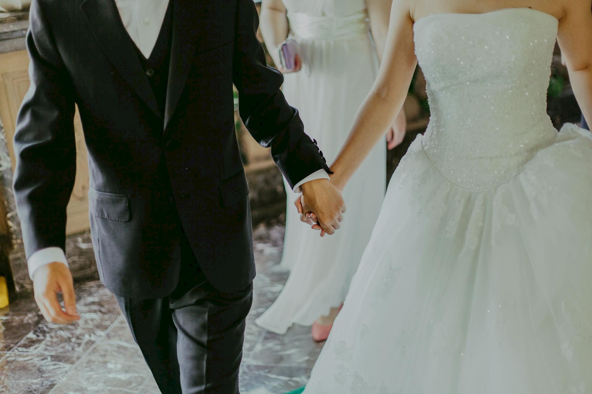 A couple dressed in wedding attire walking hand in hand; a bride in a white gown and a groom in a suit, holding hands.