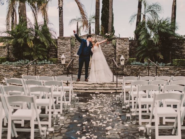 A wedding ceremony moment: a couple in formal attire kiss on a stone stage, surrounded by palms and white chairs.