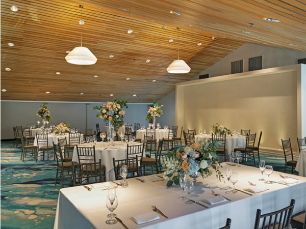 A wedding reception with banquet tables, white tablecloths, floral centerpieces, and hanging pendant lights in a hall.