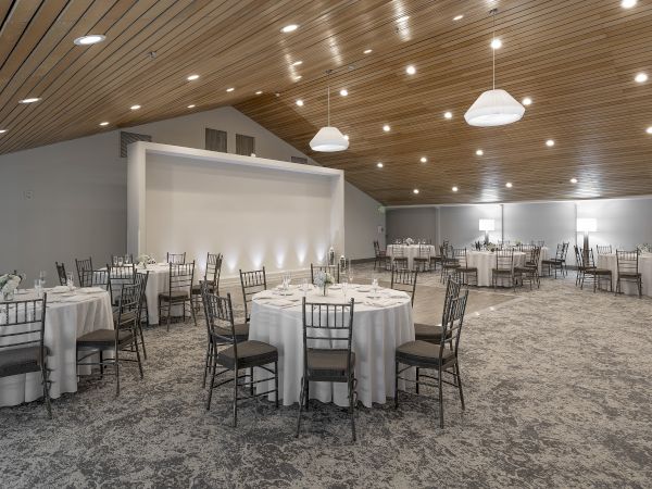 A modern banquet hall set with tables and chairs, white linens, and hanging pendant lights under a wooden ceiling.