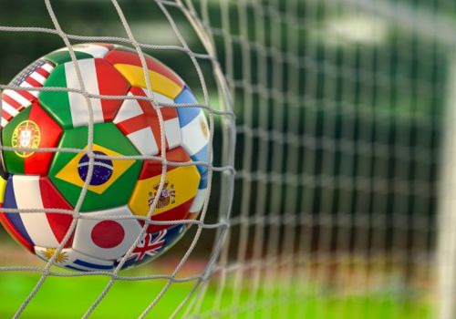 A colorful soccer ball with multiple national flags sits behind a goal net, with the stadium fence blurred in the background.
