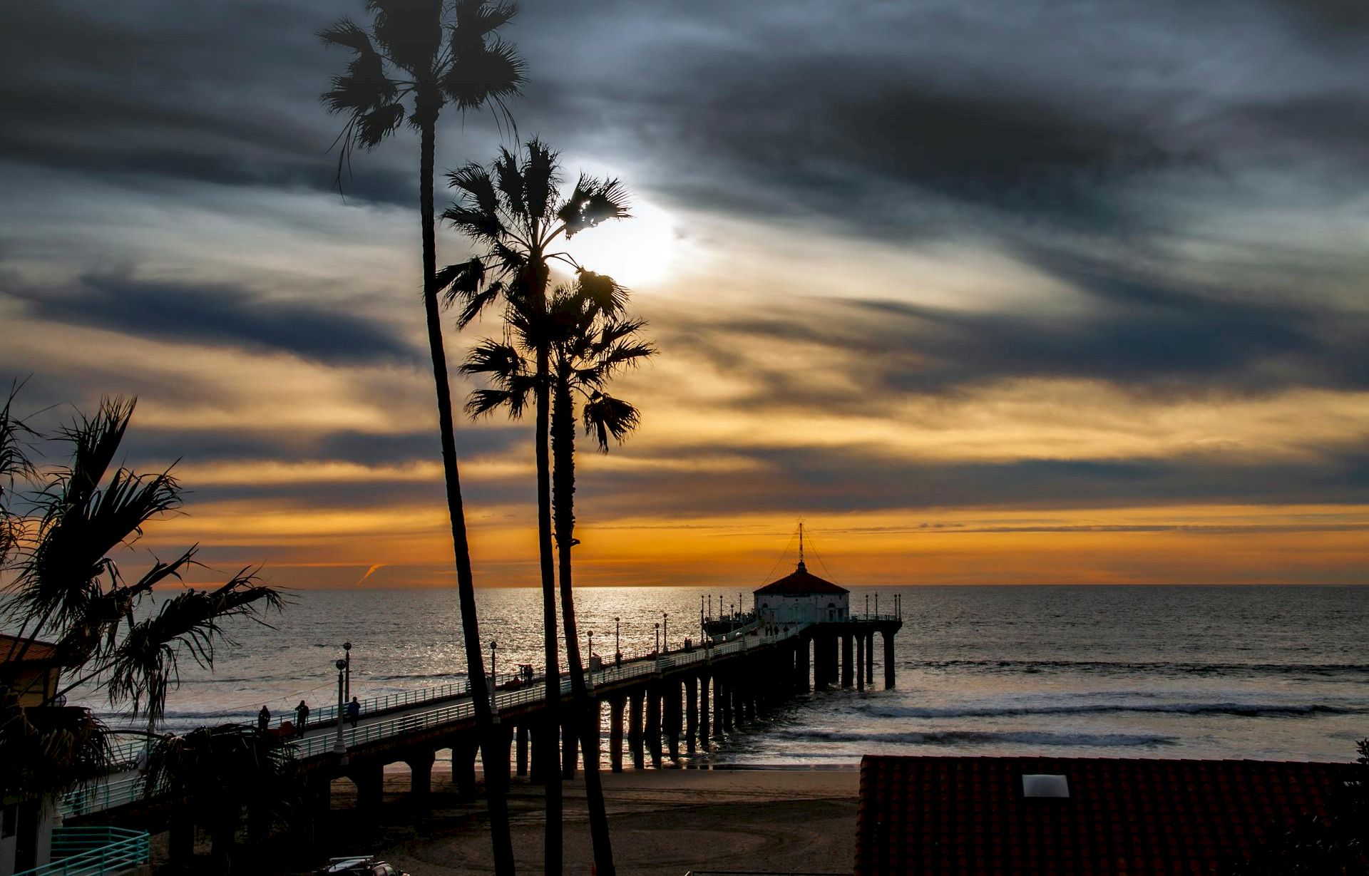 A seaside pier with a gazebo juts into the ocean as silhouetted palm trees frame a dramatic sunset under stormy clouds.
