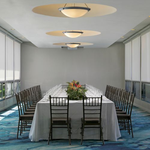 A banquet room setup with a long dining table, chairs, a floral centerpiece, and recessed ceiling lamps glowing softly.