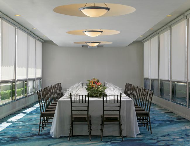 A banquet room setup with a long dining table, chairs, a floral centerpiece, and recessed ceiling lamps glowing softly.