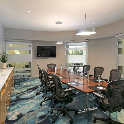 Modern office conference room with long table, swivel chairs, TV, ceiling lights, and blinds-covered windows.