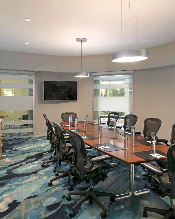 Modern office conference room with long table, swivel chairs, TV, ceiling lights, and blinds-covered windows.