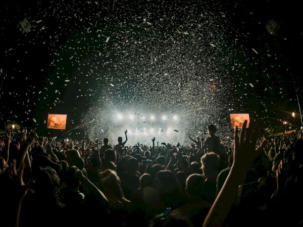 A crowded concert at night with bright stage lights, confetti in the air, and silhouettes of cheering fans raising their hands.