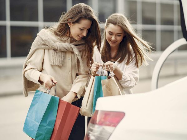Two young women with shopping bags loading purchases into a car trunk, smiling and chatting after a shopping trip.