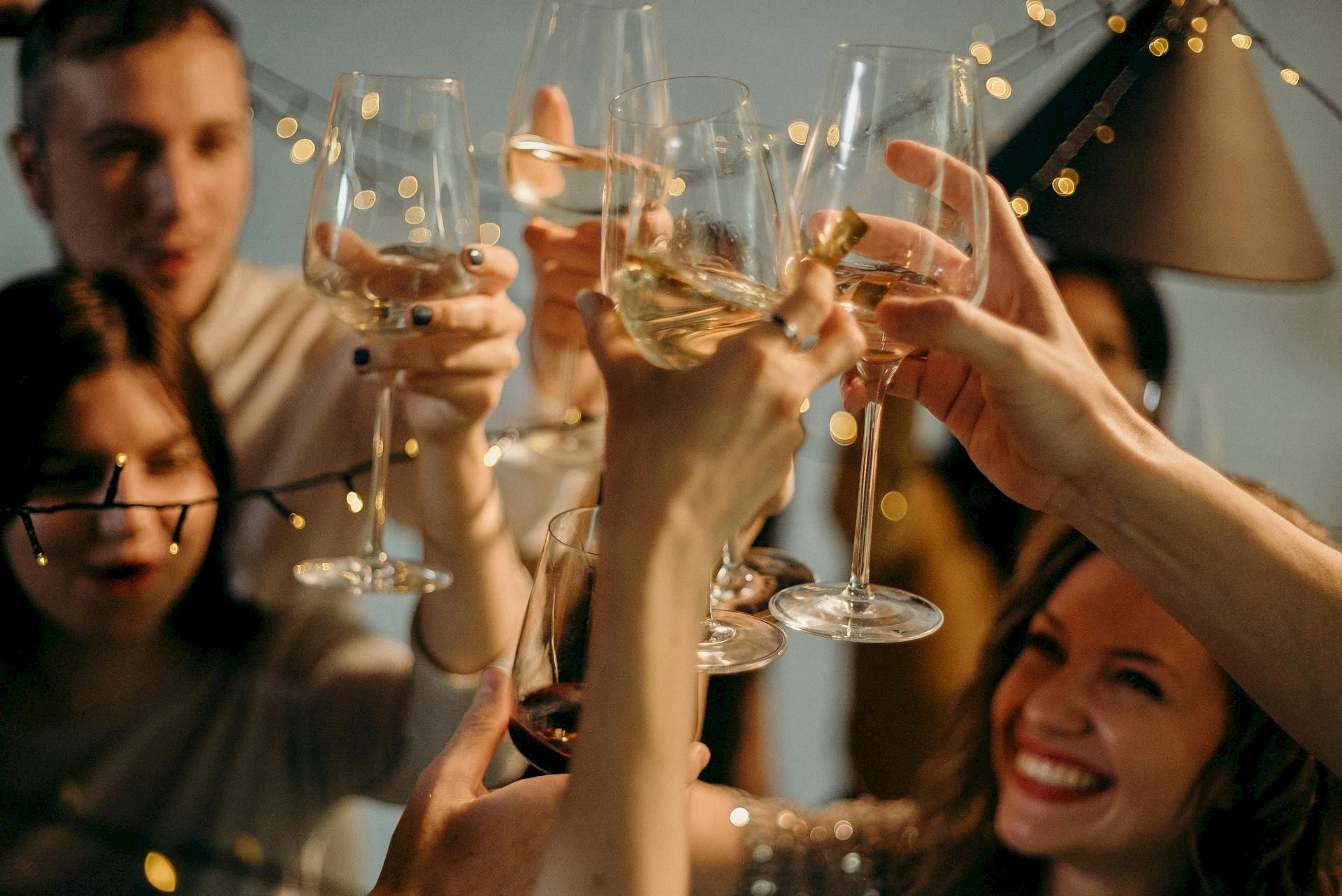 Friends clink wine glasses at a celebration, with golden bokeh lights floating overhead.