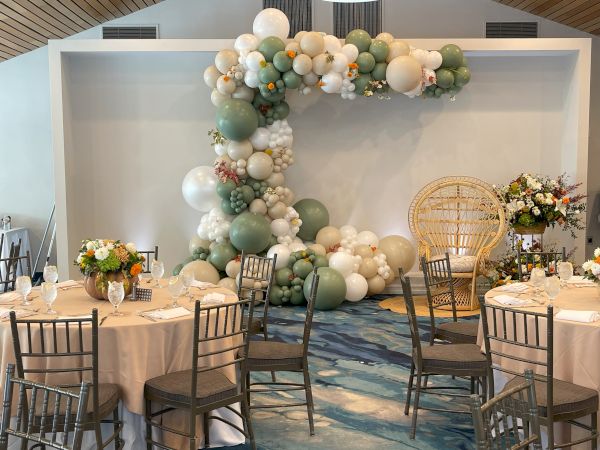 A banquet reception setup with a pastel balloon arch, floral centerpieces, and chairs around a decorated banquet table.