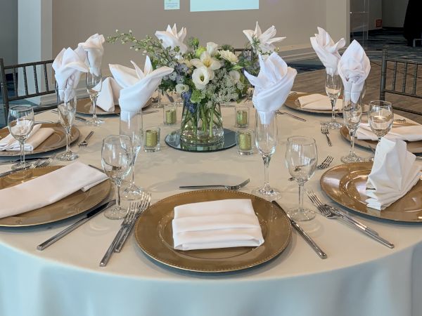 An elegant banquet table set with white napkins, gold plates, crystal glassware, and a centerpiece bouquet of white flowers.