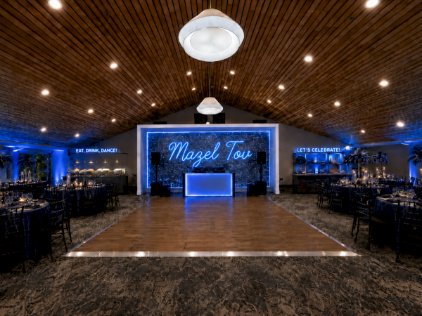 An illuminated event hall with candlelit tables, blue lighting, a central dance stage, and a neon &ldquo;Magel Tou&rdquo; sign above.
