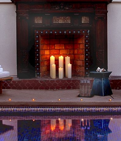 A cozy poolside spa setup with two white lounge chairs, neatly folded towels, a glowing fireplace with candles, and a small table by the side, all reflected in the pool.