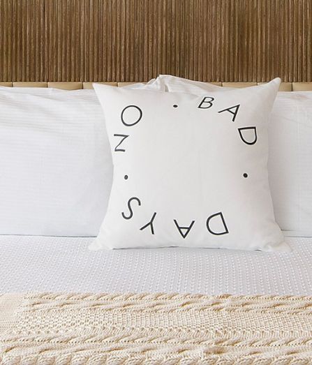 A neatly made bed with white linens, two matching lamps, a textured throw at foot, and a decorative pillow reading &ldquo;DAYS&rdquo; on a wood-paneled headboard.
