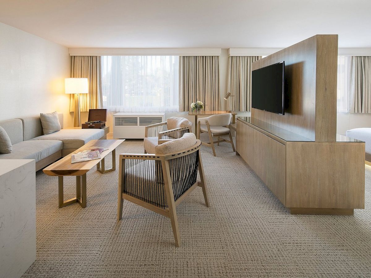 Modern hotel lounge with sofas, a coffee table, chairs, a TV on a wood console, and big curtained windows.