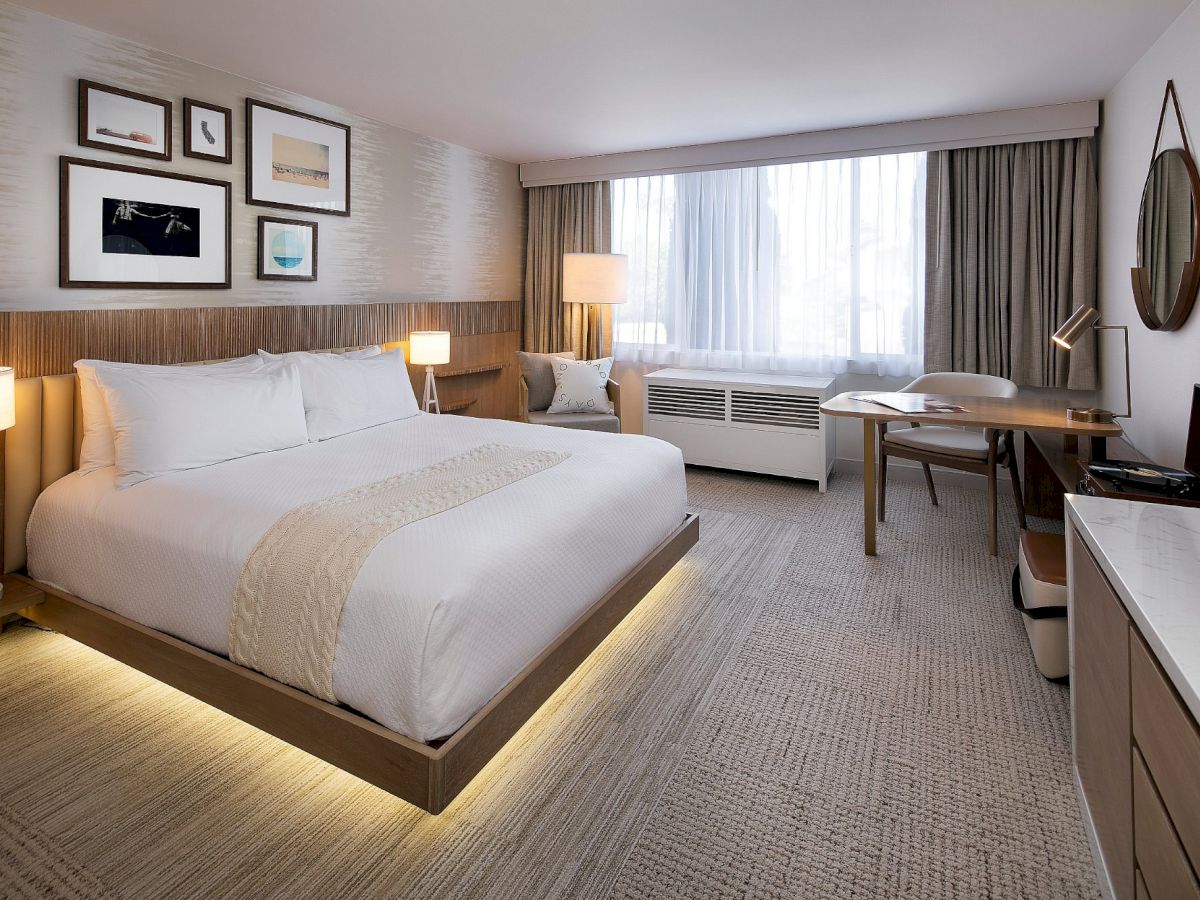 A modern hotel room with a king-size bed, bedside lamps, framed wall art, a desk, and large window curtains.