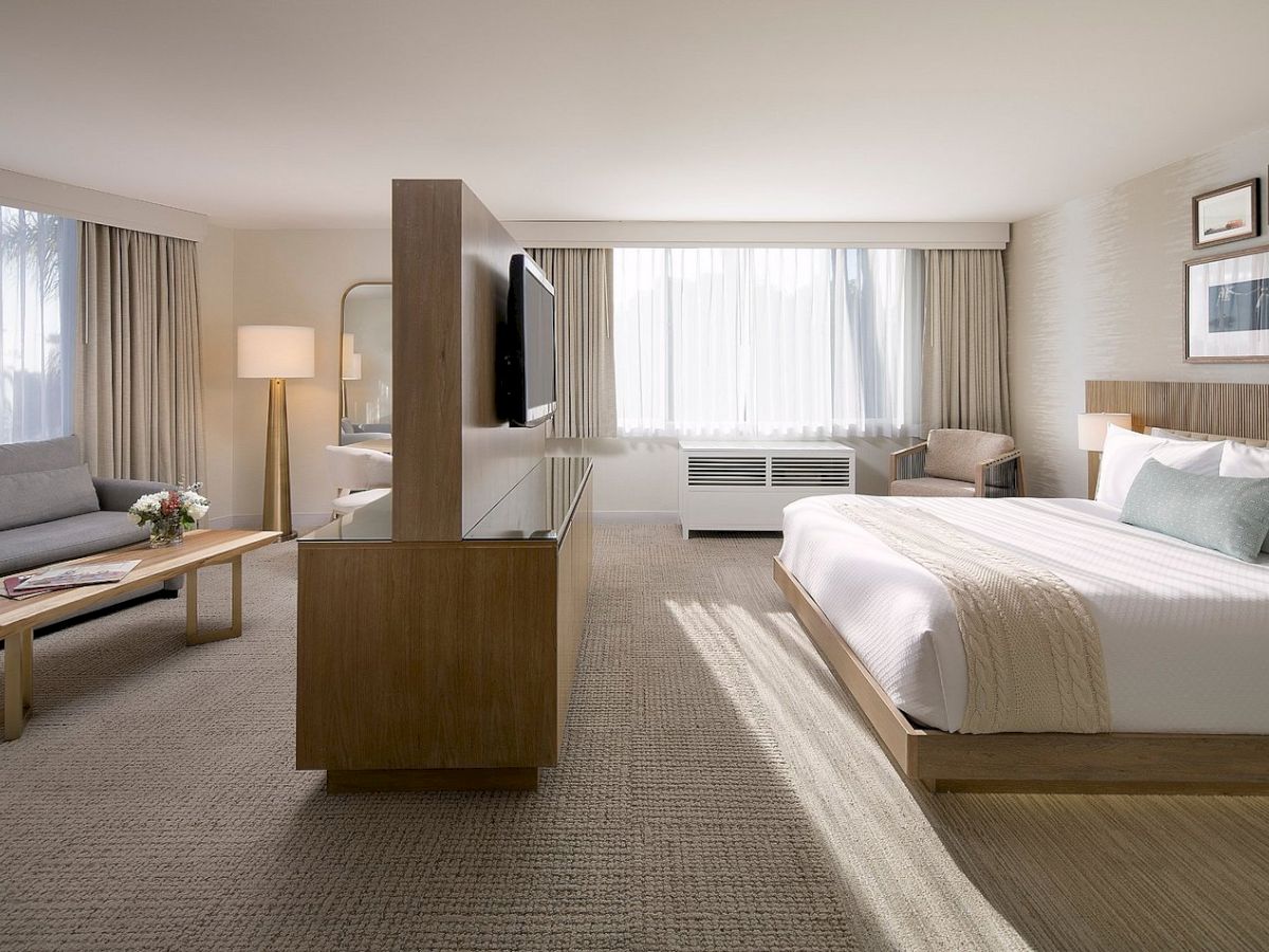 A modern hotel room with a bed, sofa, TV stand, lamps, curtains, and framed art in a bright, neutral space.