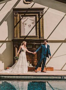 A bride and groom in formalwear stand by a tiled fireplace wall beside a swimming pool, under sunny striped shadows.