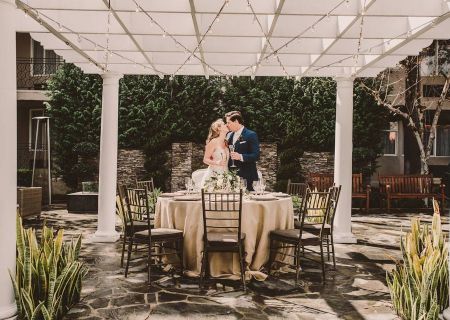 An outdoor wedding reception: a couple seated at a set table under a white canopy with string lights and greenery around them.
