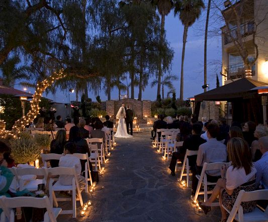 An outdoor wedding ceremony at dusk with rows of guests, candles along an aisle, palm trees, and a bride walking forward.