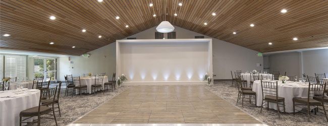 A bright banquet hall with round tables, chairs, a central dance floor, and a raised stage under recessed ceiling lights.
