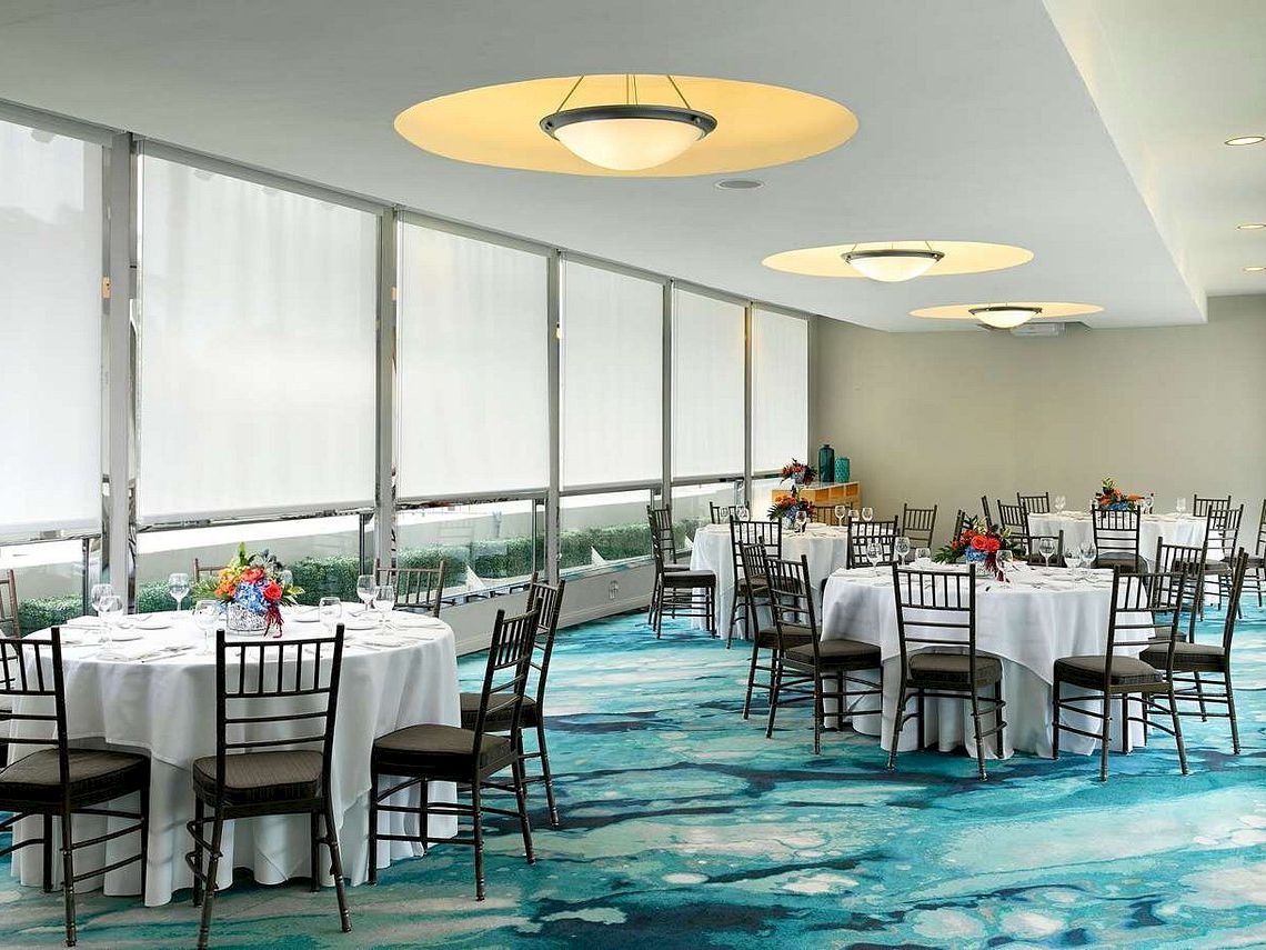A bright banquet hall with round tables, floral centerpieces, and black chairs by large windows, with a blue patterned carpet.