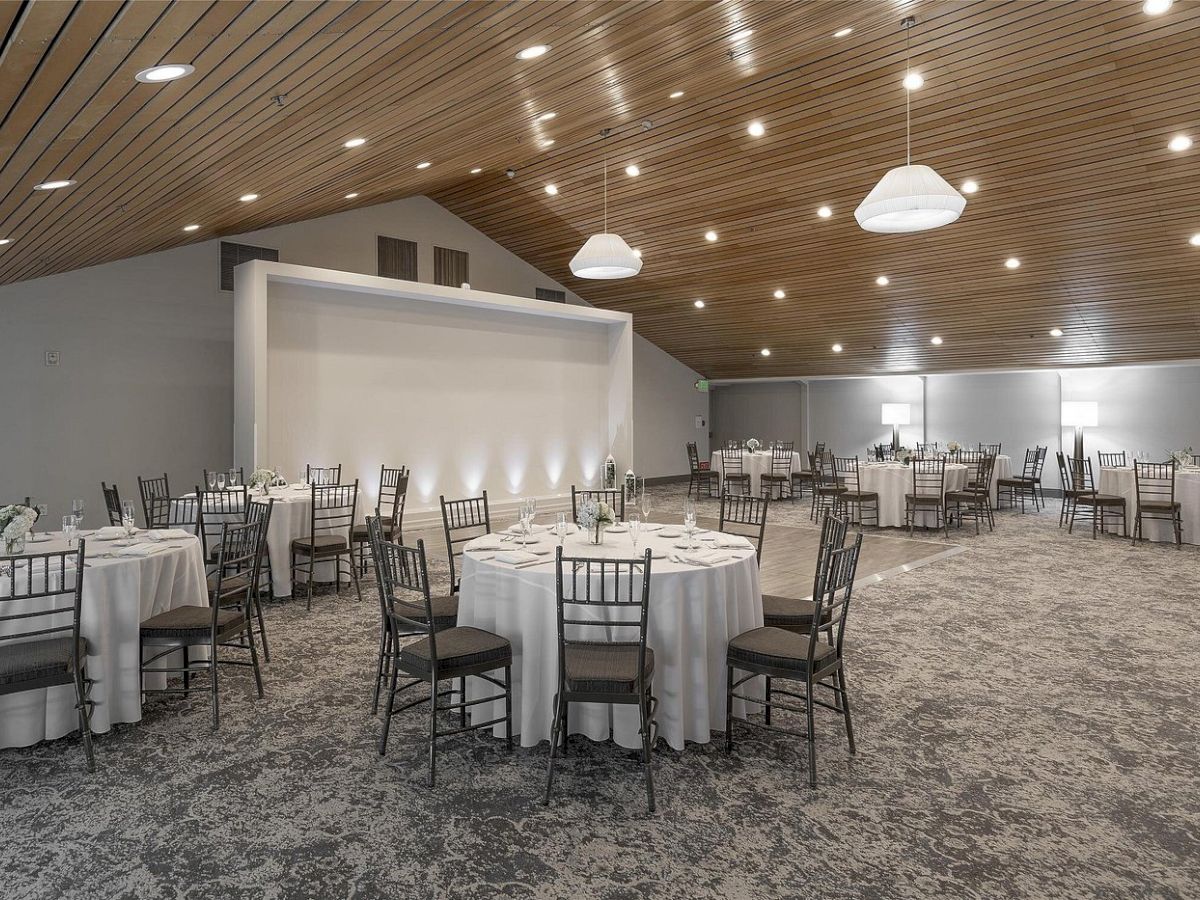 A banquet hall set for a formal event: round tables with white linens, black chairs, and warm ceiling lights.