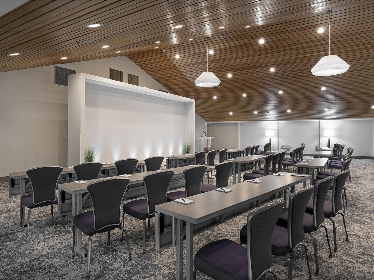 A modern conference room with long tables, black chairs, overhead pendant lights, and a large presentation screen area.