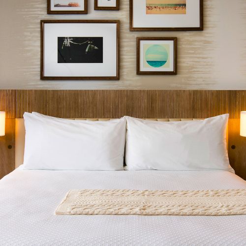 A neatly made hotel bedroom with a white bed, two bedside lamps, and framed seaside photos on the wall.