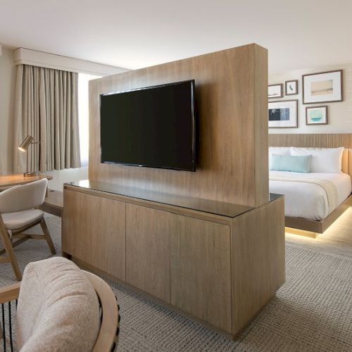 A modern hotel suite with a wall-mounted TV on a wooden divider facing a lounge area and a bed, warm lighting, and framed pictures on the wall.