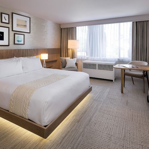 A modern hotel room with a bed, bedside lamps, desk, TV, framed wall art, and a large window with curtains.