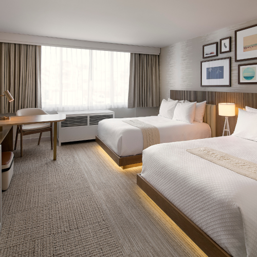 A modern hotel room with two beds, a desk and TV, and large windows with curtains plus framed wall art.