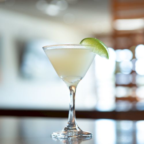 A close-up of a sparkling cocktail glass with a lemon twist on a bar, bokeh lights in the background, and a shallow depth of field.