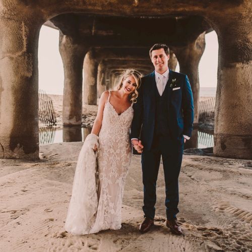 A newlywed couple stands on a sandy beach under a pier, holding hands; the bride wears a lace gown and the groom a dark suit. End.