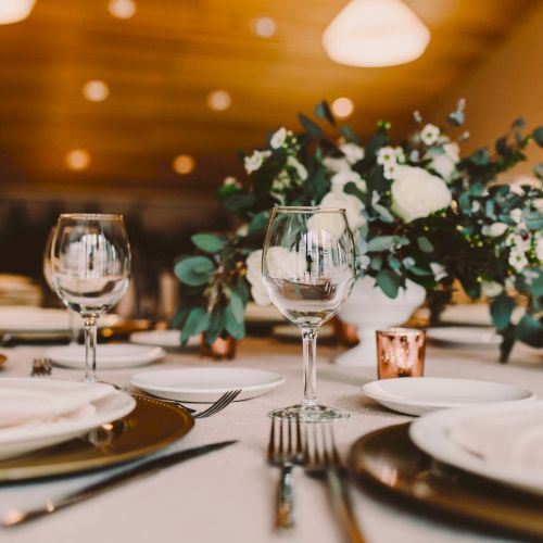 A formal dining table set with white plates, silverware, wine glasses, and a green floral centerpiece, under warm ambient lighting.
