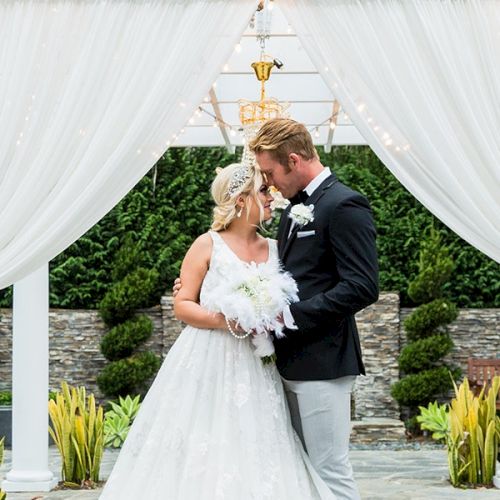 A wedding couple in white and black stands under a white draped arch, surrounded by greenery and palm frond decor.